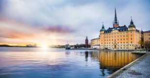 Solnedgång över vattnet i Stockholm med historiska byggnader och en kyrkspira i förgrunden.
