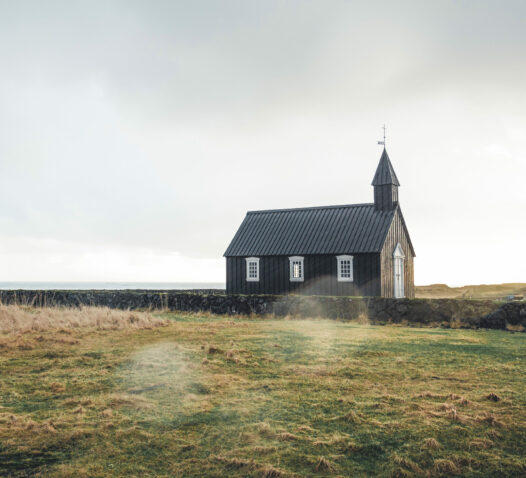 En liten svart kyrka står på en öppen äng under en mulen himmel.
