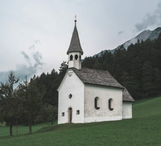 En liten vit kyrka med ett spetsigt torn står på en grön kulle framför skogsklädda berg.