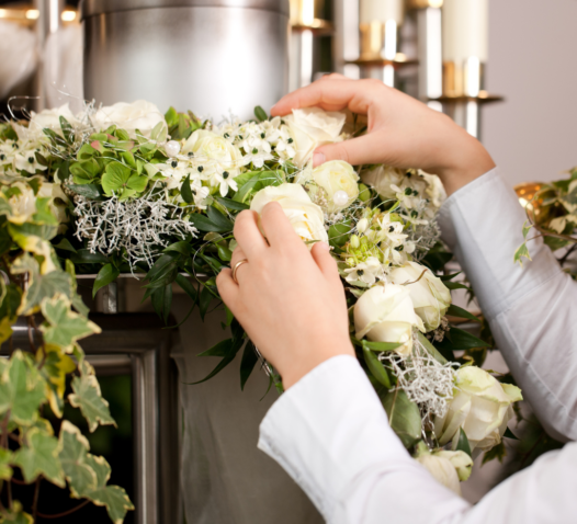 Händer arrangerar vita rosor och gröna blad runt en urna.
