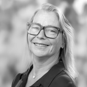 En kvinna med glasögon ler mot kameran, bakgrunden är suddig.