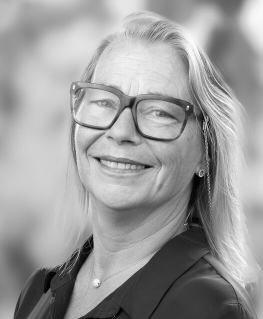 En kvinna med glasögon ler mot kameran, bakgrunden är suddig.