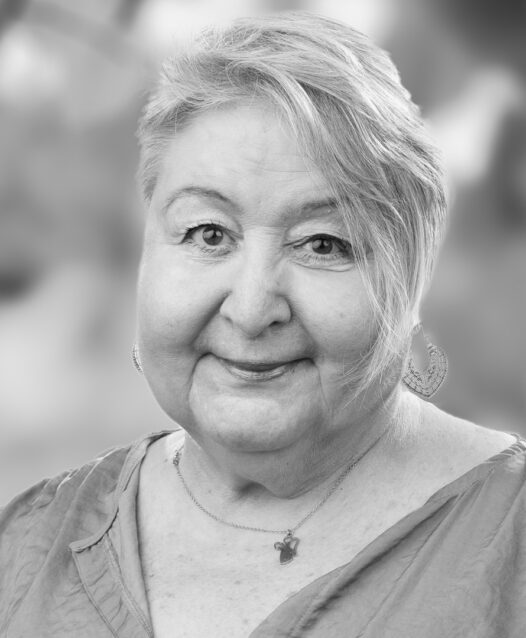 En äldre kvinna ler vänligt mot kameran, suddig bakgrund.