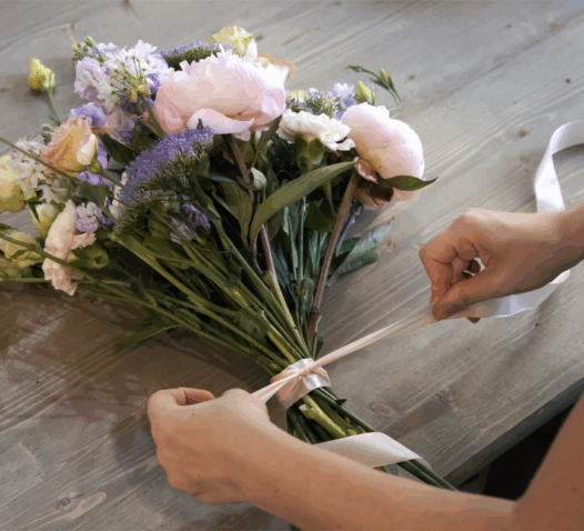 Händer binder ett band runt en bukett med blommor på ett träbord.
