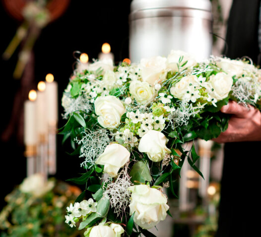 En person håller en stor blomsterarrangemang med vita rosor och gröna blad framför tända ljus.